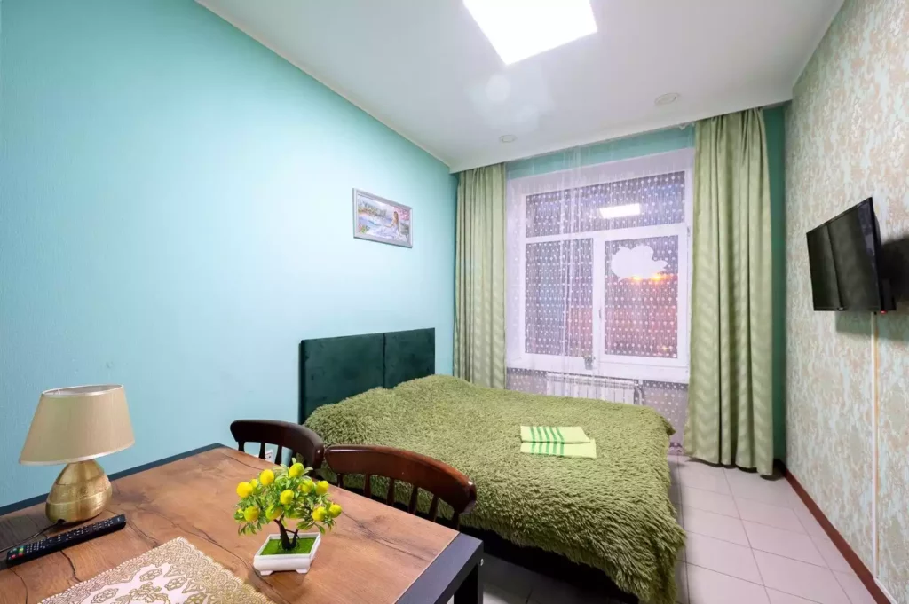 Номер с двуспальной кроватью столом, ТВ и окном. Зеленый плед и занавески. 